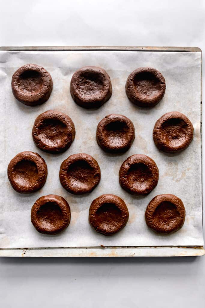 Chocolate cookies shaped in 1 inch balls with thumbprints on a lined baking tray