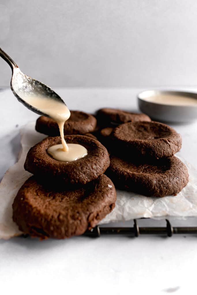 Tahini being poured into dark chocolate thumbprint cookies on a wire rack