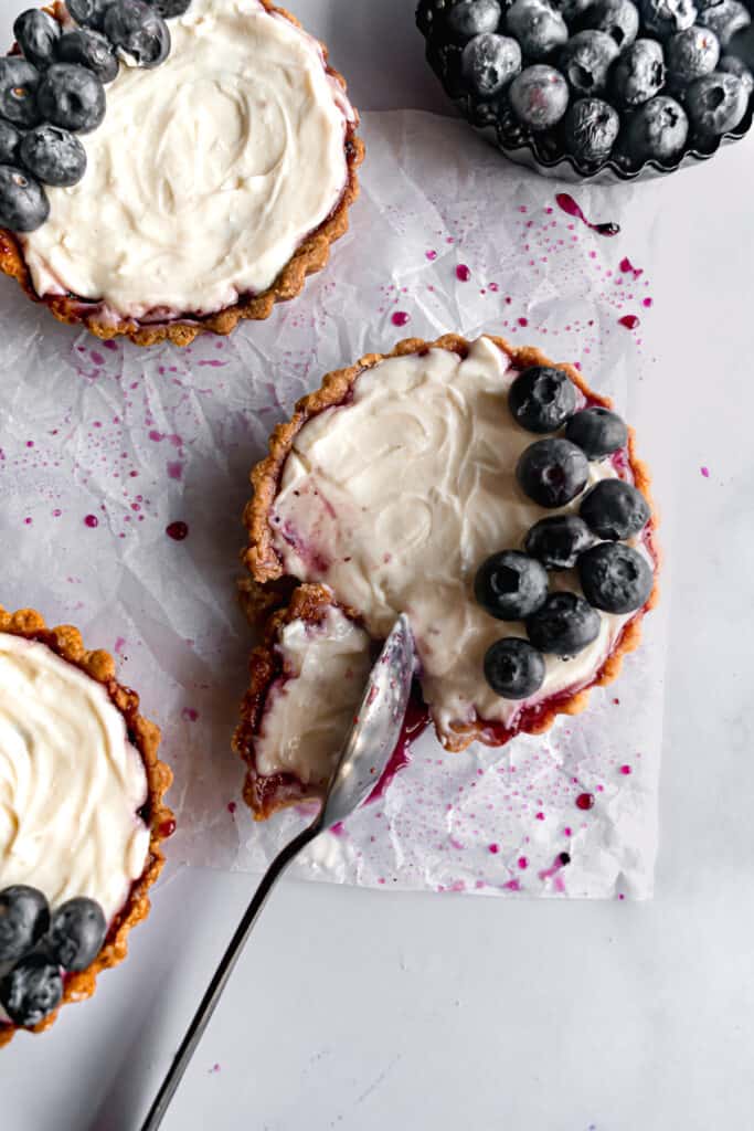 Tartlets with a cheesecake filling and blueberries with one bite in a spoon on a white parchment paper