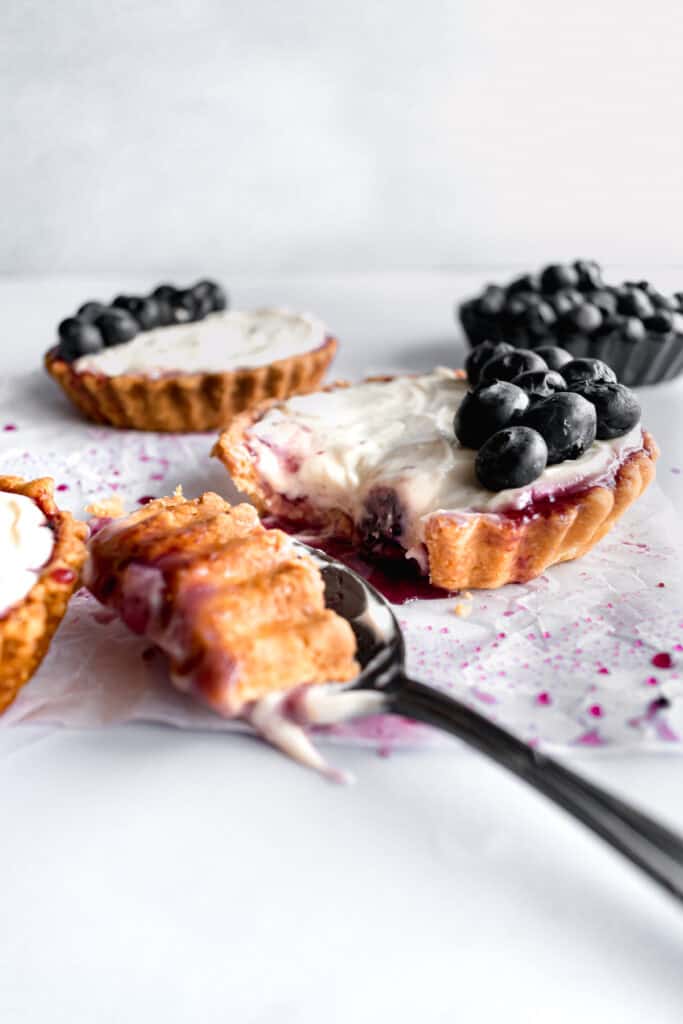 Mini blueberry tarts with cheesecake filling and one bite in a spoon on a white parchment paper