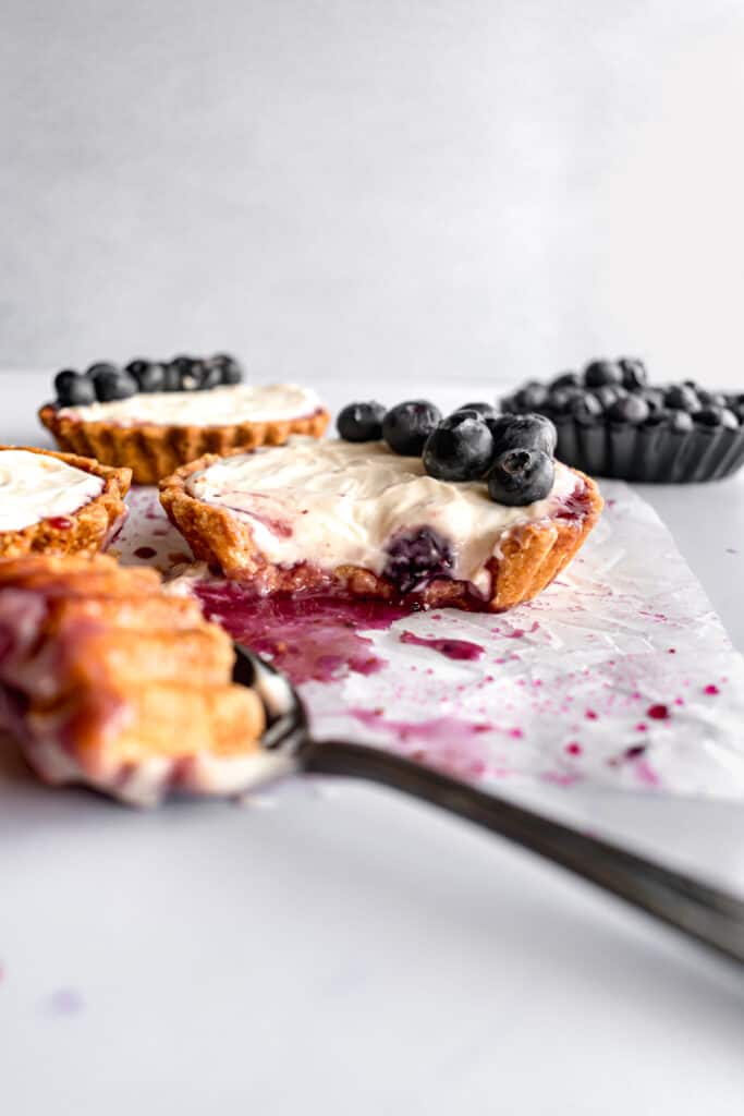 Mini blueberry tarts with cheesecake filling and one bite in a spoon on a white parchment paper