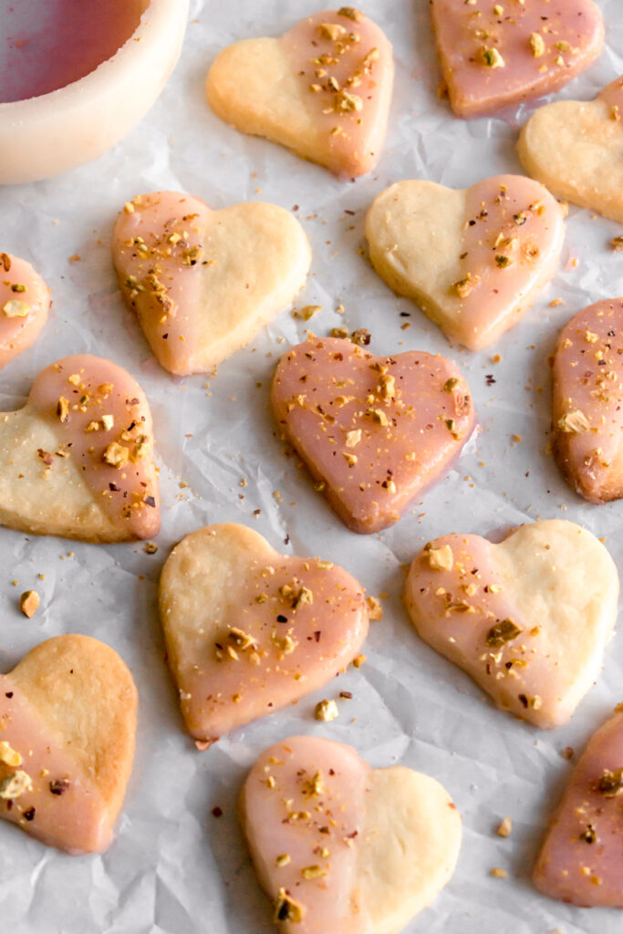 heart shaped shortbread cardamom cookies dipped in with rose flavoured glaze and topped with crushed pistachios on a parchment paper