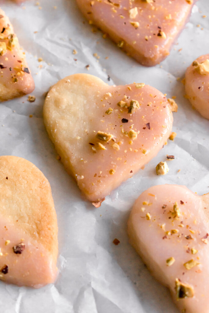 heart shaped shortbread cardamom cookies dipped in with rose flavoured glaze and topped with crushed pistachios on a parchment paper