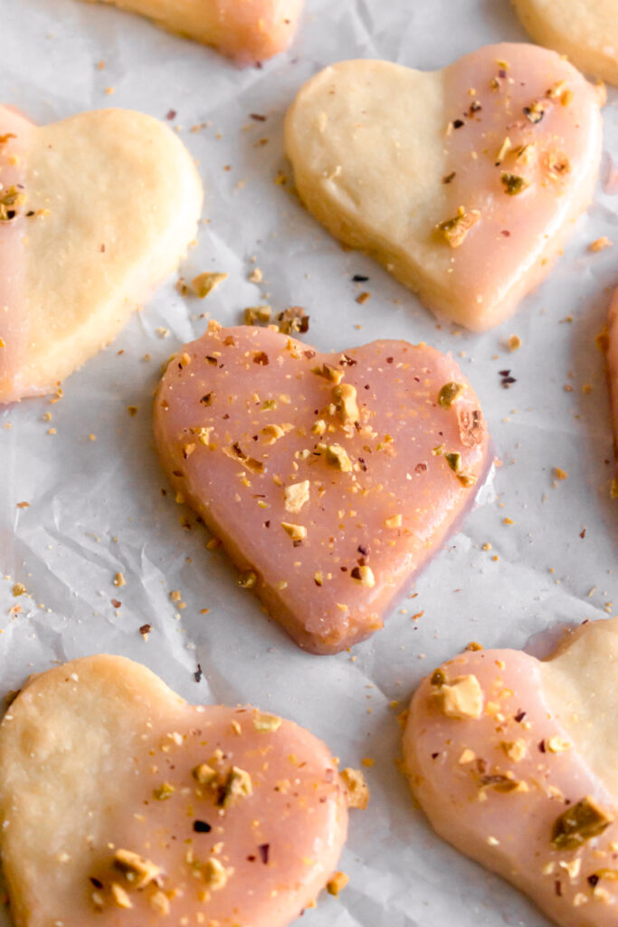 heart shaped shortbread cardamom cookies dipped in with rose flavoured glaze and topped with crushed pistachios on a parchment paper