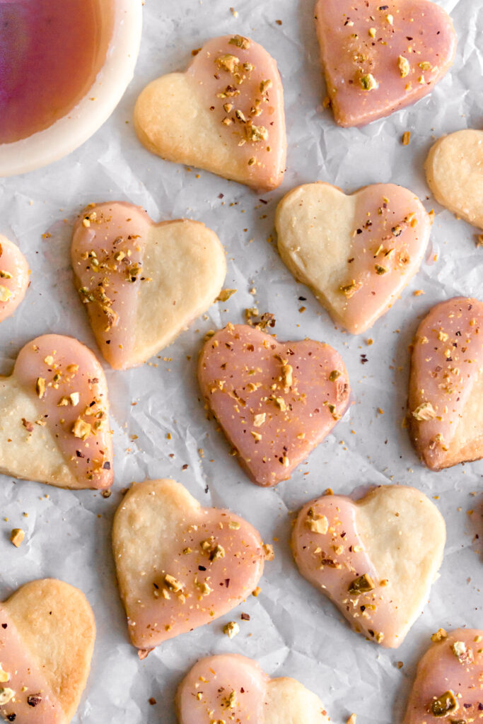 heart shaped shortbread cardamom cookies dipped in with rose flavoured glaze and topped with crushed pistachios on a parchment paper